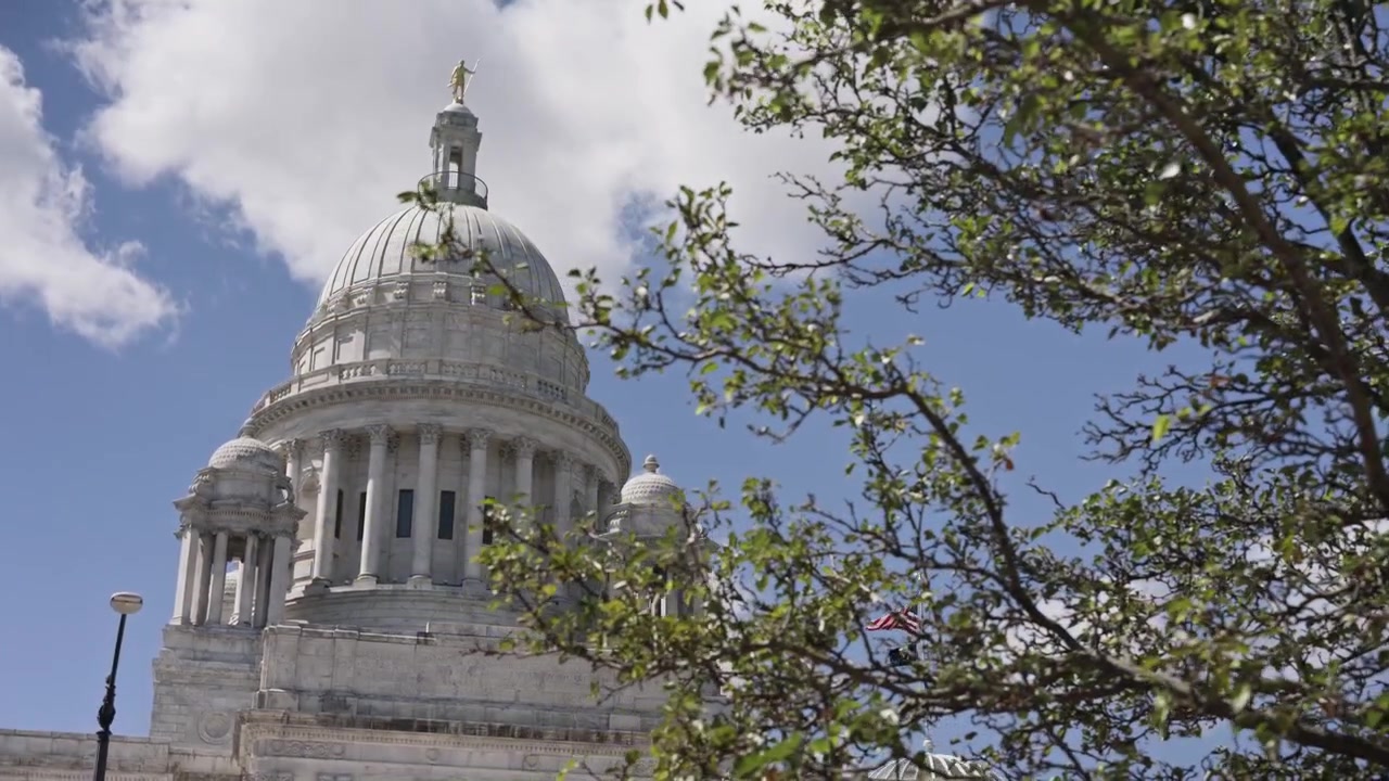 Plano cinematográfico com dolly para a esquerda do Capitólio de Rhode Island com uma árvore frondosa em primeiro plano num dia de verão ensolarado em Providence, Rhode Island.