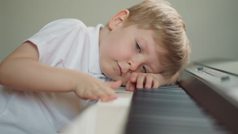 niño lindo descansa la cabeza en el piano moviendo los dedos a lo largo de las teclas perezosamente