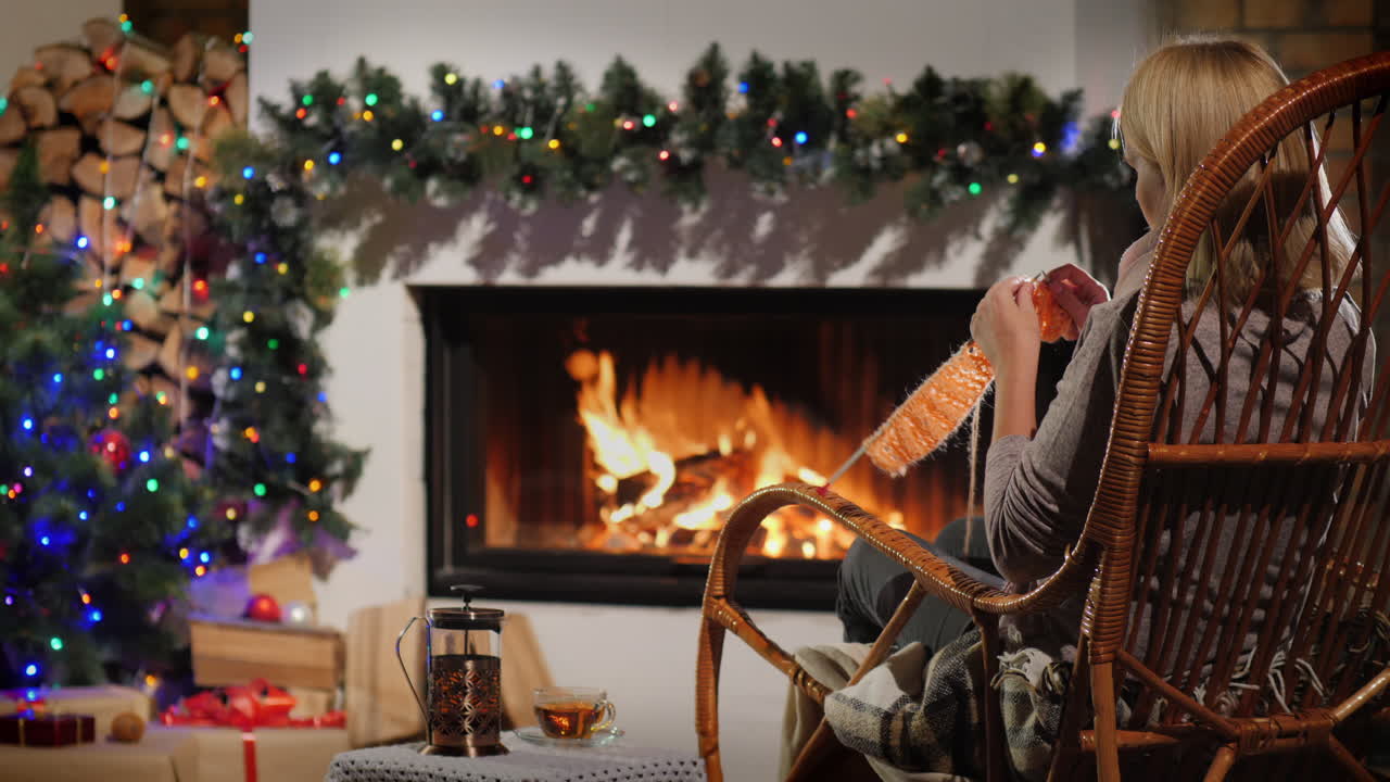 겨울 저녁에 휴식 - 크리스마스를 위해 장식된 벽난로 옆에 앉아 뜨개질을 하는 여성