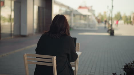 rear view of woman working on laptop outdoors with her hair covering her face, decorative black pillar nearby, blurred urban background featuring modern buildings, and figure walking in the distant