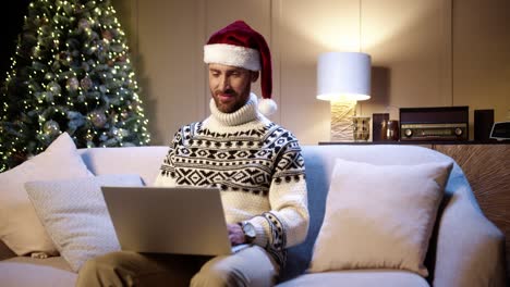 Portrait-Of-Happy-Young-Man-Wearing-Santa-Hat-Sitting-In-Decorated-Room-Near-Glowing-Christmas-Tree-Tapping-And-Browsing-On-Laptop