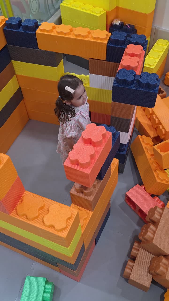 A five-year-old girl concentrates as she lifts a large, soft building block, engaging in creative and constructive play at a colorful indoor playground or kids cafe.