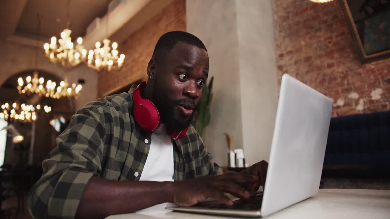 A black person with a beard in red headphones works on a laptop in a cafe, rejoices in success and raises his hands up as a sign of victory. Emotions of success at your favorite job