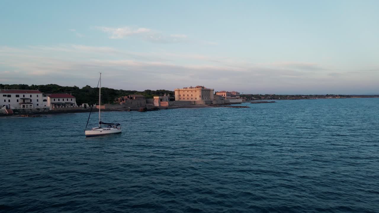 un dron se dirige hacia un antiguo castillo en la costa de italia mientras veleros y pescadores adornan el mar.