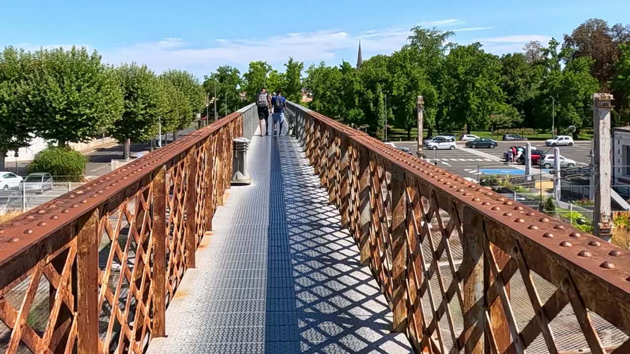 A rusty pedestrian bridge crosses over railway tracks, with nearby buildings and greenery visible.
