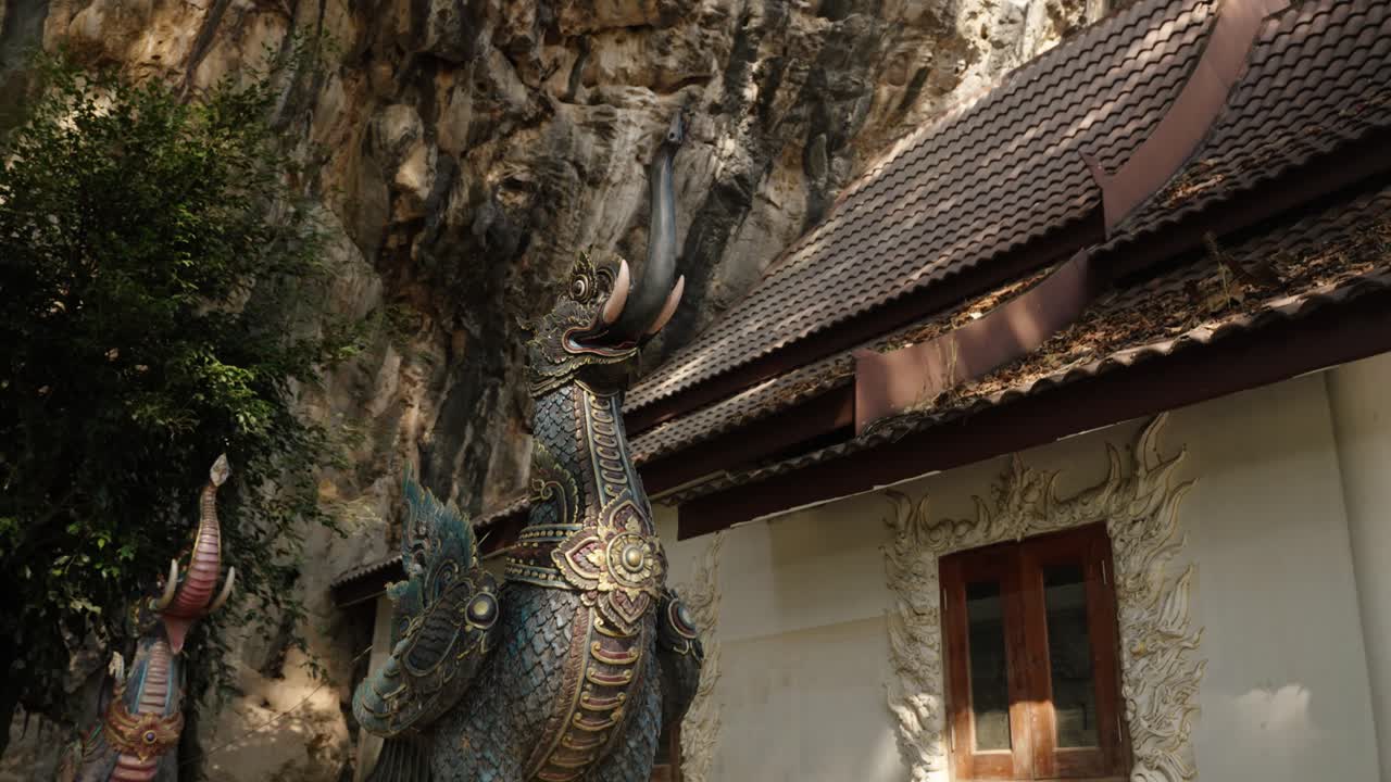 Ornate temple guardian statue stands beside a cliffside hall in Wat Tham Phra