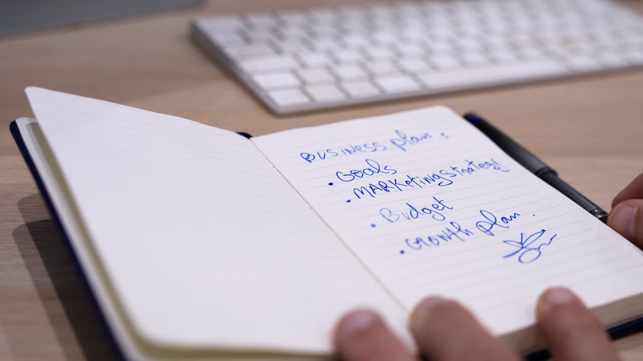 Close-up of a hand signing a completed business plan in a notebook on an office desk beside a computer keyboard, symbolizing agreement, approval, strategy execution, and professional decision making