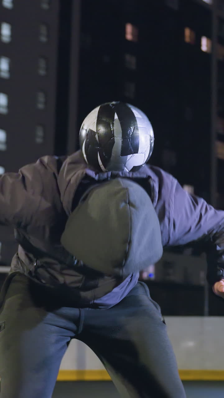 el deportista lanza hábilmente la pelota, la equilibra en la espalda, la empuja hacia arriba y la atrapa durante la práctica nocturna en un campo rodeado de edificios residenciales iluminados y farolas brillantes