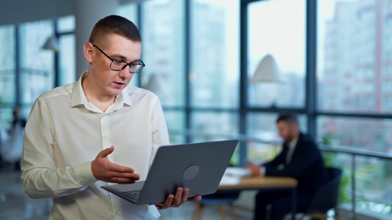 Male manager with laptop in one hand stands in office. Young man describes something on computer. Man in suit in blur at backdrop.