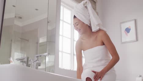 video of smiling biracial woman with towel on hair sitting on bathtub in bathroom