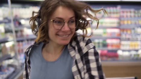 At-the-supermarket:-happy-young-girl-funny-dancing-between-shelves-in-supermarket.-Curly-girl-wearing-black-and-white-plaid-shirt-with-headphones-on-head-listening-to-her-favorite-music.-Slow-motion.-Close-up