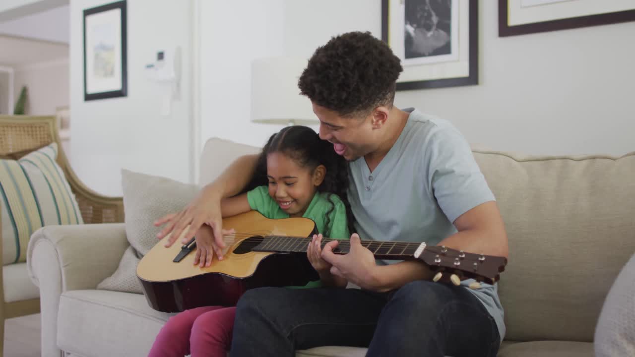padre y hija biraciales felices sentados en el sofá tocando la guitarra