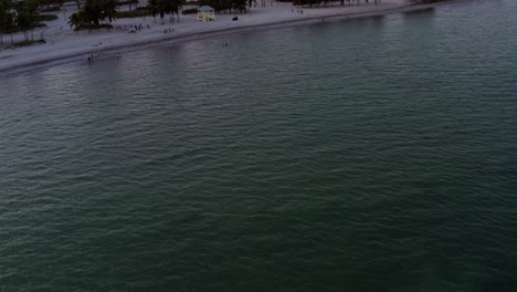 Tilt-up-dolly-in-aerial-drone-shot-revealing-the-tropical-beach-surrounded-by-palm-trees-on-Crandon-Park-in-Key-Biscayne-with-the-skyline-of-Miami,-Florida-in-the-distance-on-a-sunny-summer-evening