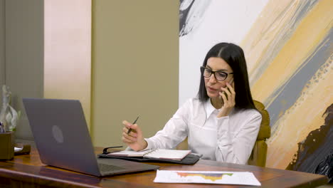 business woman talking on mobile phone and writing something in the agenda while sitting at desk in the office