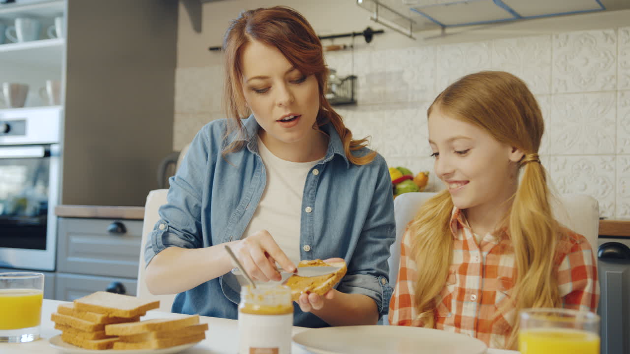 아름다운 어머니가 그녀의 예쁜 금발 십대 소녀에게 멋진 아 ⁇ 한 부 ⁇ 에서  ⁇ 에 땅콩 버터를 퍼뜨리는 방법을 보여줍니다. 초상화 촬영. 안쪽