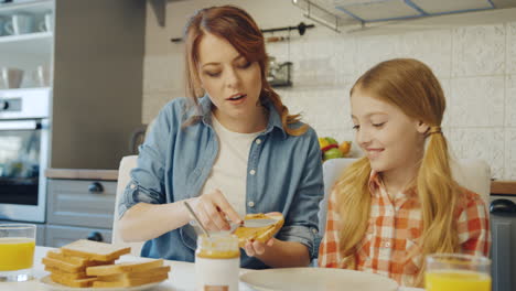 Beautiful-mother-showing-to-her-pretty-blonde-teen-girl-how-to-spread-the-peanut-butter-on-the-bread-in-the-nice-cozy-kitchen.-Portrait-shot.-Inside