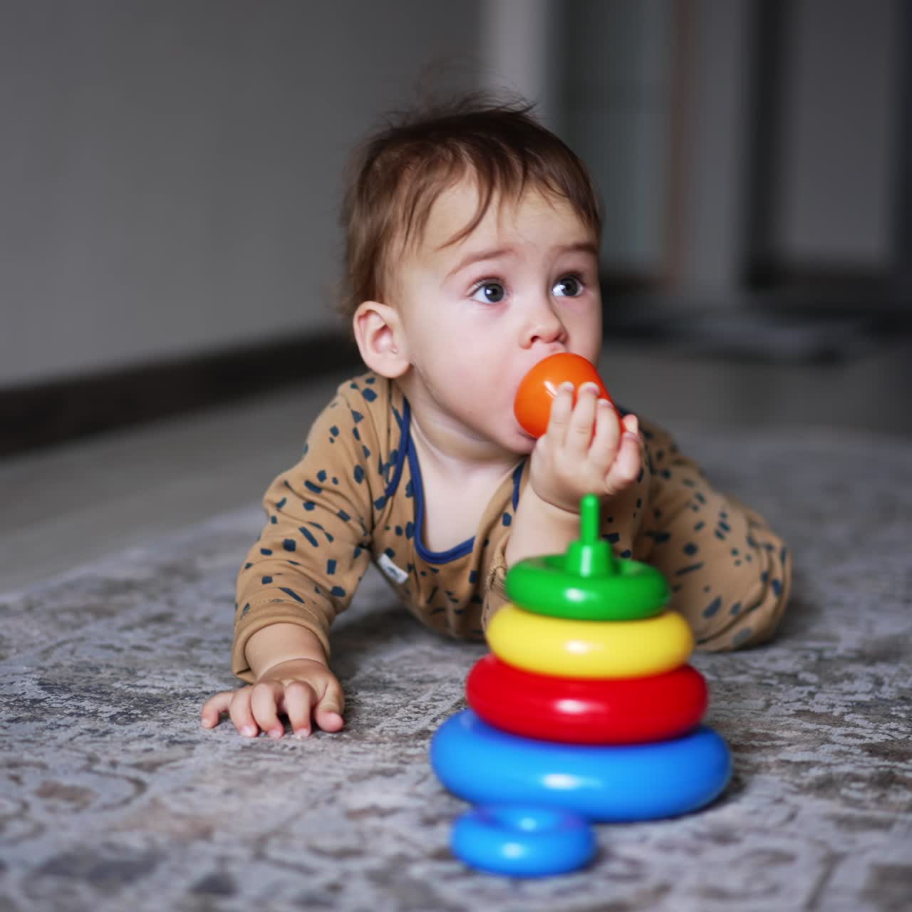 Baby boy in brown romper lies on the carpet indoors. Adorable one-year old child playing with a toy pyramid. Blurred backdrop
