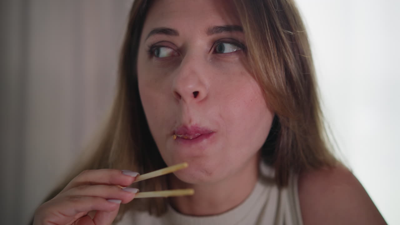 Close up of lady using chopsticks to savour flavorful meat sushi roll indoors, eyes focused on food, slow motion bite showing enjoyment, natural expression with soft lighting and neutral background
