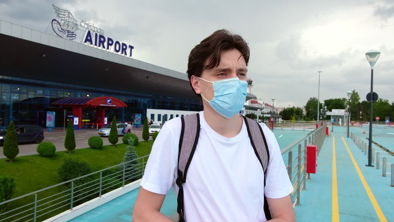 CHISINAU, MOLDOVA - JUNE 19, 2020: Man with a white t-shirt, backpack and surgical mask waiting for the flight in front of the airport. Traveling during Corona Virus pandemic outbreak. Chisinau, Moldova