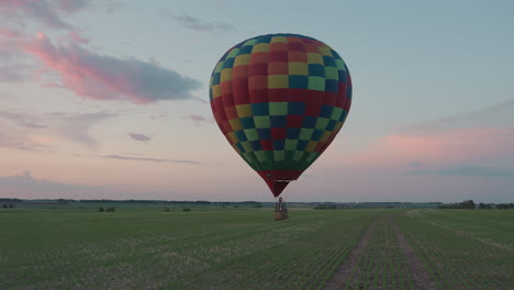 passengers in hot air balloon drifting over vast green farmland at sunset burner flame illuminating wicker basket while distant car headlights sparkle under soft pastel evening sky