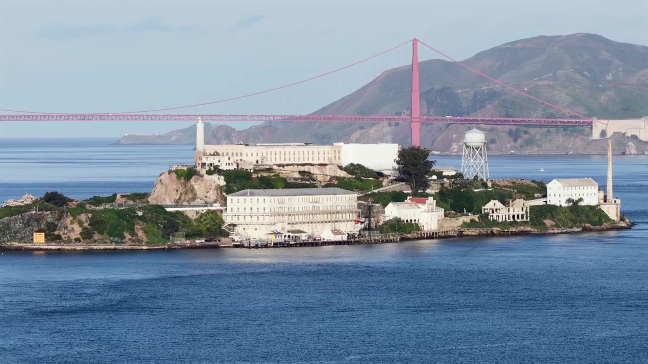 アメリカ合衆国カリフォルニア州サンフランシスコ湾に浮かぶアルカトラズ島。歴史的な刑務所建築、灯台、給水塔があり、ゴールデンゲートブリッジと海岸の丘を背景に穏やかな景色の光に包まれている。航空ドローンからの眺め。