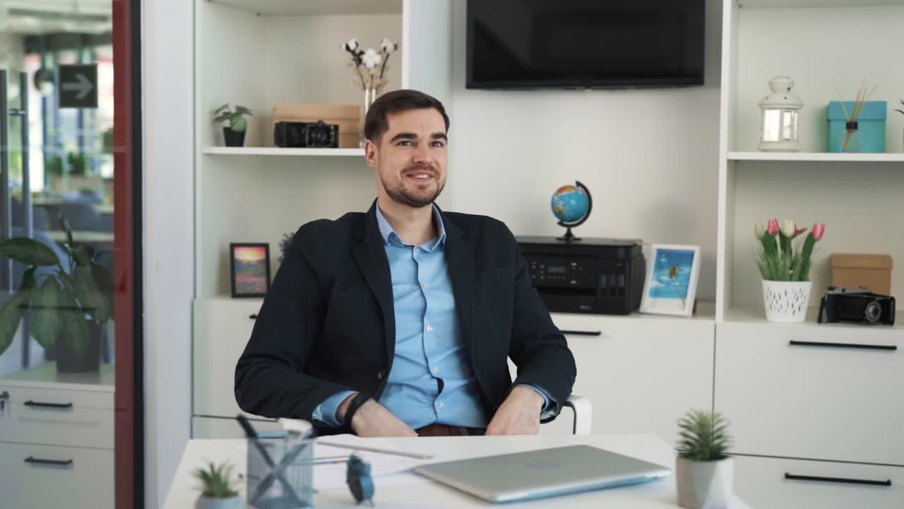 The young man in a suit sits at a desk in the office and thinks captively about something, envisioning a happy future and his success