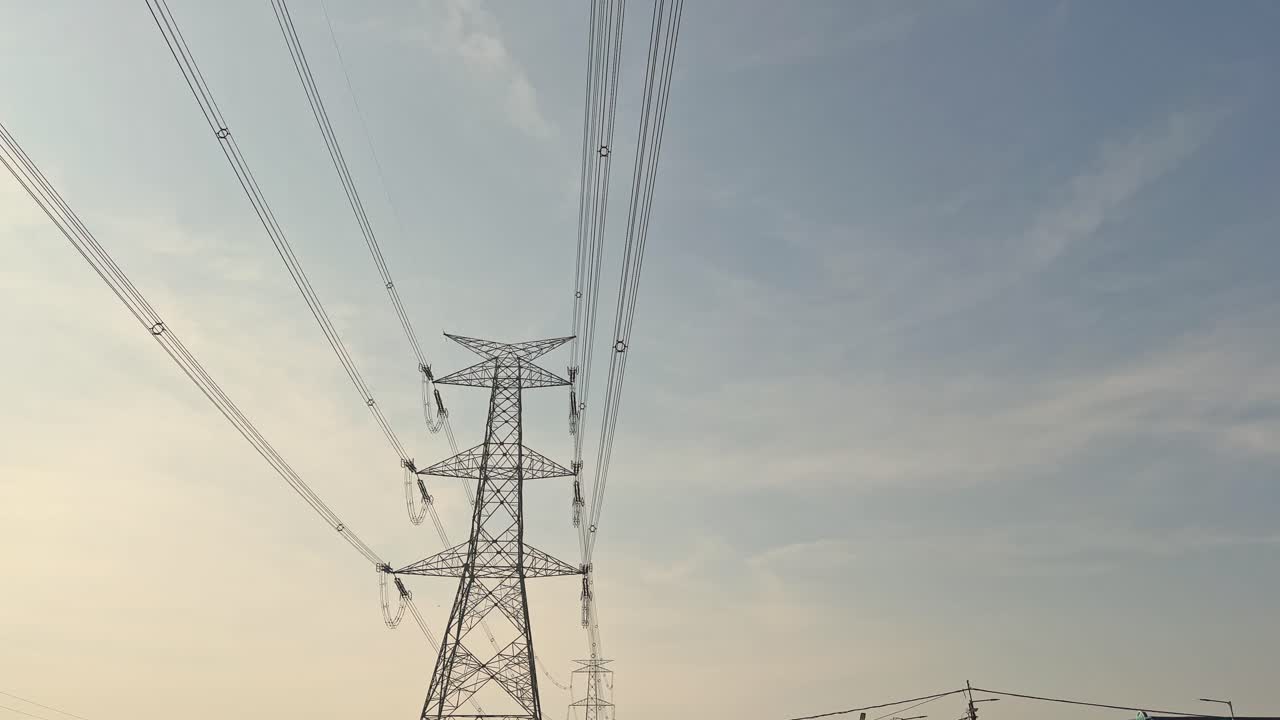 Tilt-up view along bundled power lines leading to a high-voltage transmission tower, steel lattice rising into open sky as conductors converge in daylight calm