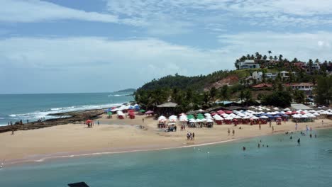 Left-trucking-aerial-drone-shot-passing-the-tropical-Tibau-do-Sul-beach-with-tourists-swimming,-enjoying-shade-under-colorful-umbrellas,-and-waves-crashing-into-rocks-in-Rio-Grande-do-Norte,-Brazil