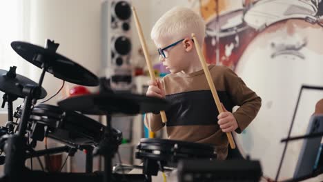 a small talented albino boy with white hair in blue round glasses plays an electronic drum musical instrument using special wooden sticks during his leisure time during the day in his room