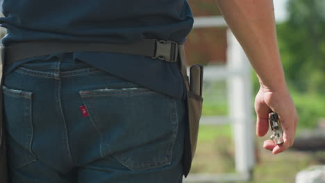 Close up rear view of adult walking outdoors holding plier in hand near garden structure, jeans and belt visible, sunlight highlighting movement and tool in casual work setting on rural plot