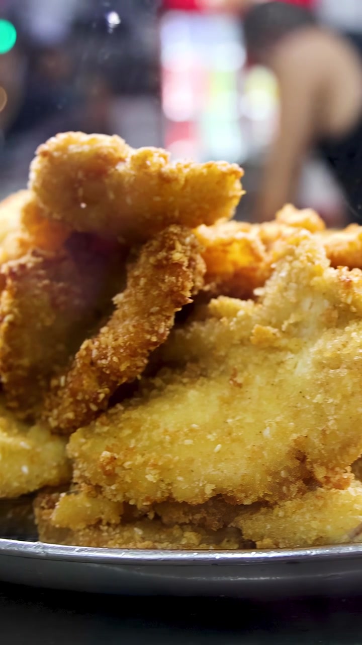 Golden breaded pork cutlets are stacked on a metal tray under bright indoor lighting, with a bustling food stall environment and slight camera pan