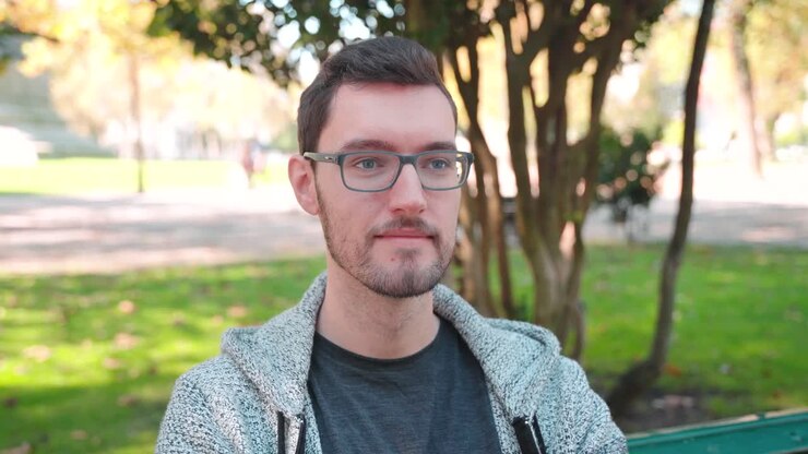 Giovane uomo con gli occhiali seduto su una panchina del parco che fissa in lontananza.