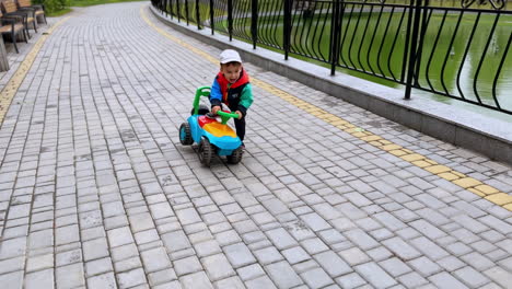 smiling caucasian kid rides his toy car in the park. toddler boy comes up to the pond fence and jumps up cheerfully.