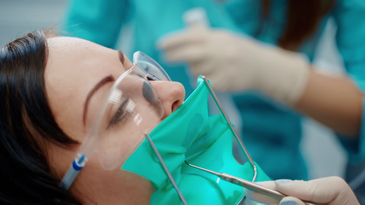 Female patient's face during tooth treatment. Young woman in protective glassses and mask in stomatology clinic. Dentist in gloves treats teeth. Close-up.