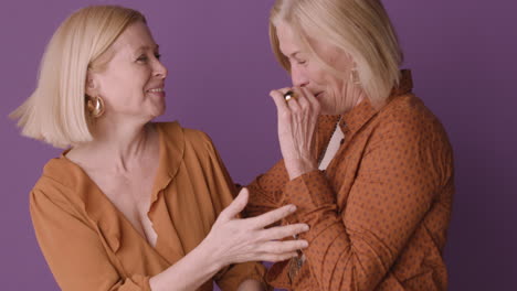 Zwei-Blonde-Reife-Frauen-In-Braunem-Hemd-Umarmen-Sich-Und-Lächeln-Auf-Violettem-Hintergrund-1