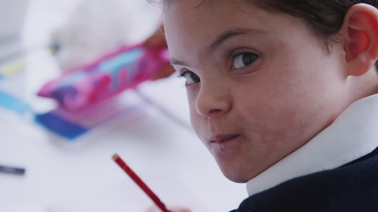 다운 증후군을 고 있는 학교 소년이 초등학교 교실에서 책상에 앉아 일하는 모습.