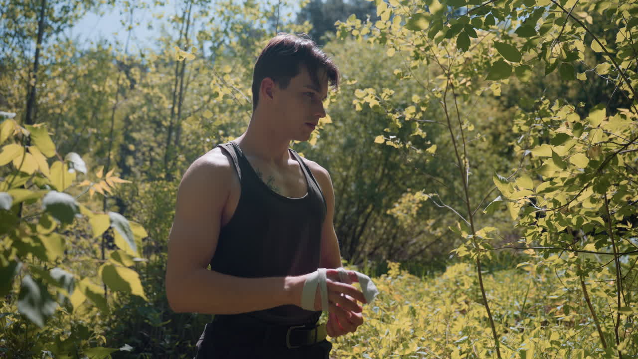 Adventurer unties white headband and rolls it while standing in middle of forest surrounded by sun filtered through leaves, tank top and belt visible, branches casting dappled light on skin