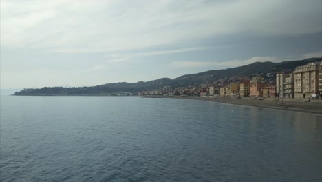 Aerial-pullback-of-vast-ocean-waters-of-the-idyllic-Italian-village-of-Varazze-revealing-three-tourists-standing-atop-stone-breakwater-looking-into-horizon