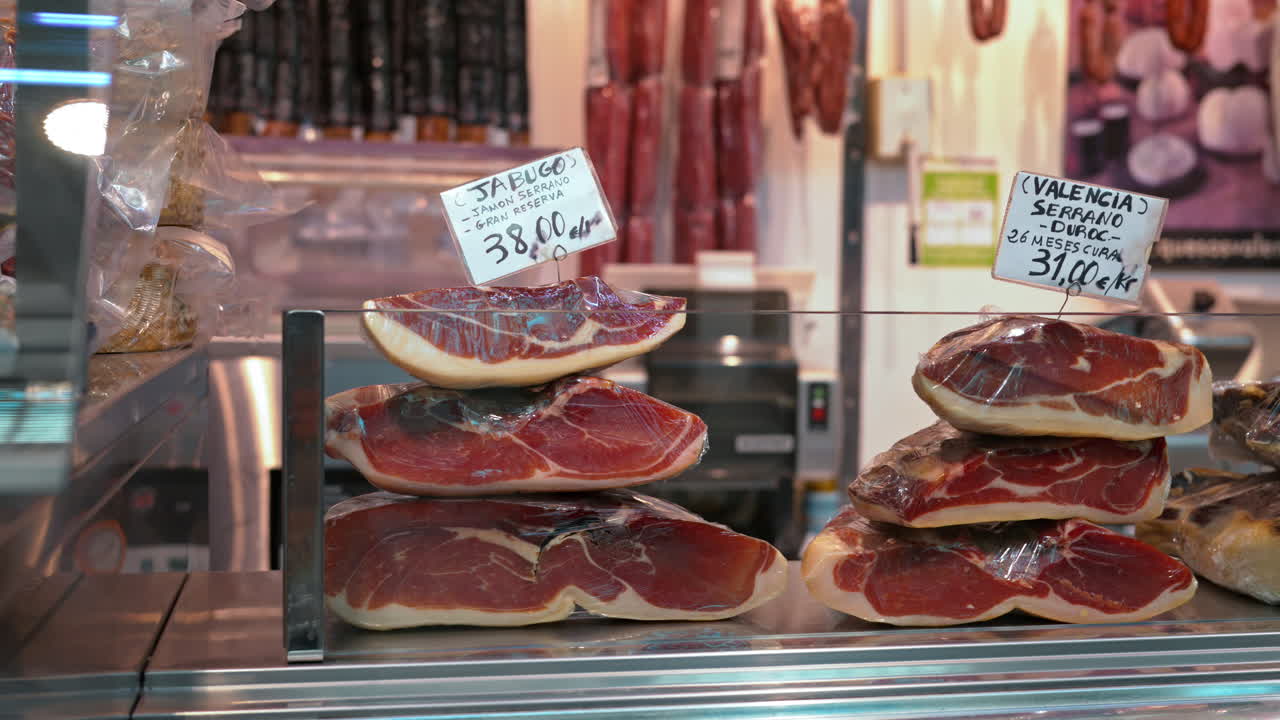Piled cuts of jamon behind a glass case with handwritten price signs and hanging sausages at a shop in Valencia, Spain