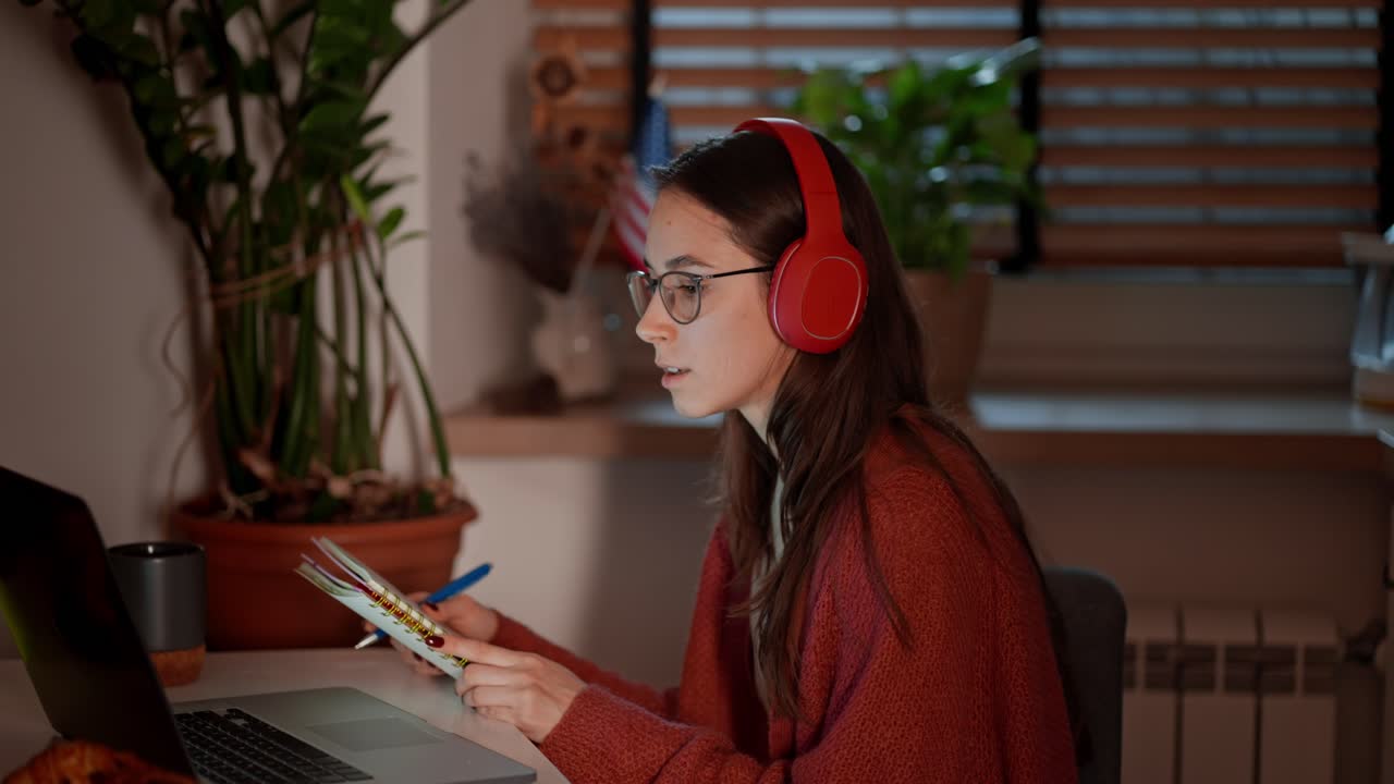 una chica morena segura con auriculares inalámbricos y gafas en un apartamento moderno en la cocina estudia lenguas extranjeras y toma notas sobre las reglas de pronunciación