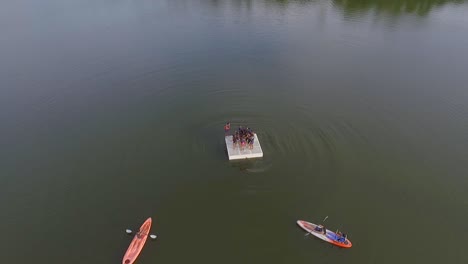 vista de drones de un grupo de niños saltando desde una plataforma de lago de campamento de verano, y dos kayaks que lo rodean