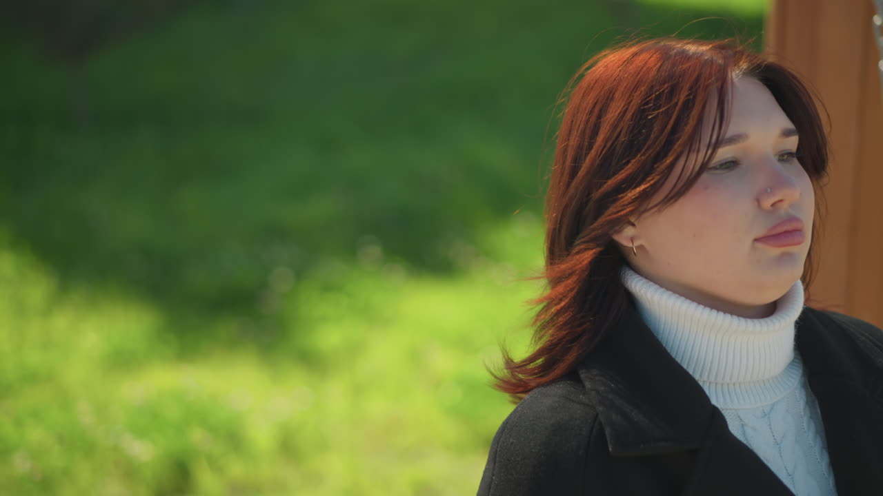 나무로 된 스에 앉아있는 여자의 클로즈업, 그녀의 머리카락은 바람에 부드럽게 불고, 빛은 울창한 초록색 가운데 그녀의 얼굴에 부드럽게 반사됩니다.