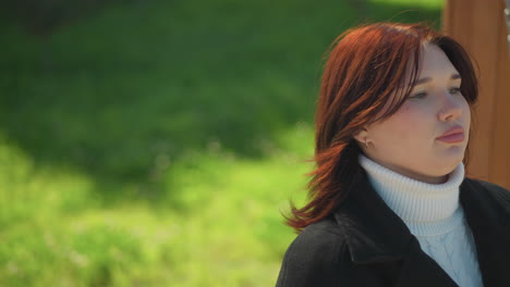 close-up of a woman sitting on a wooden swing, her hair gently blowing in the wind, with sunlight reflecting softly on her face amidst lush greenery