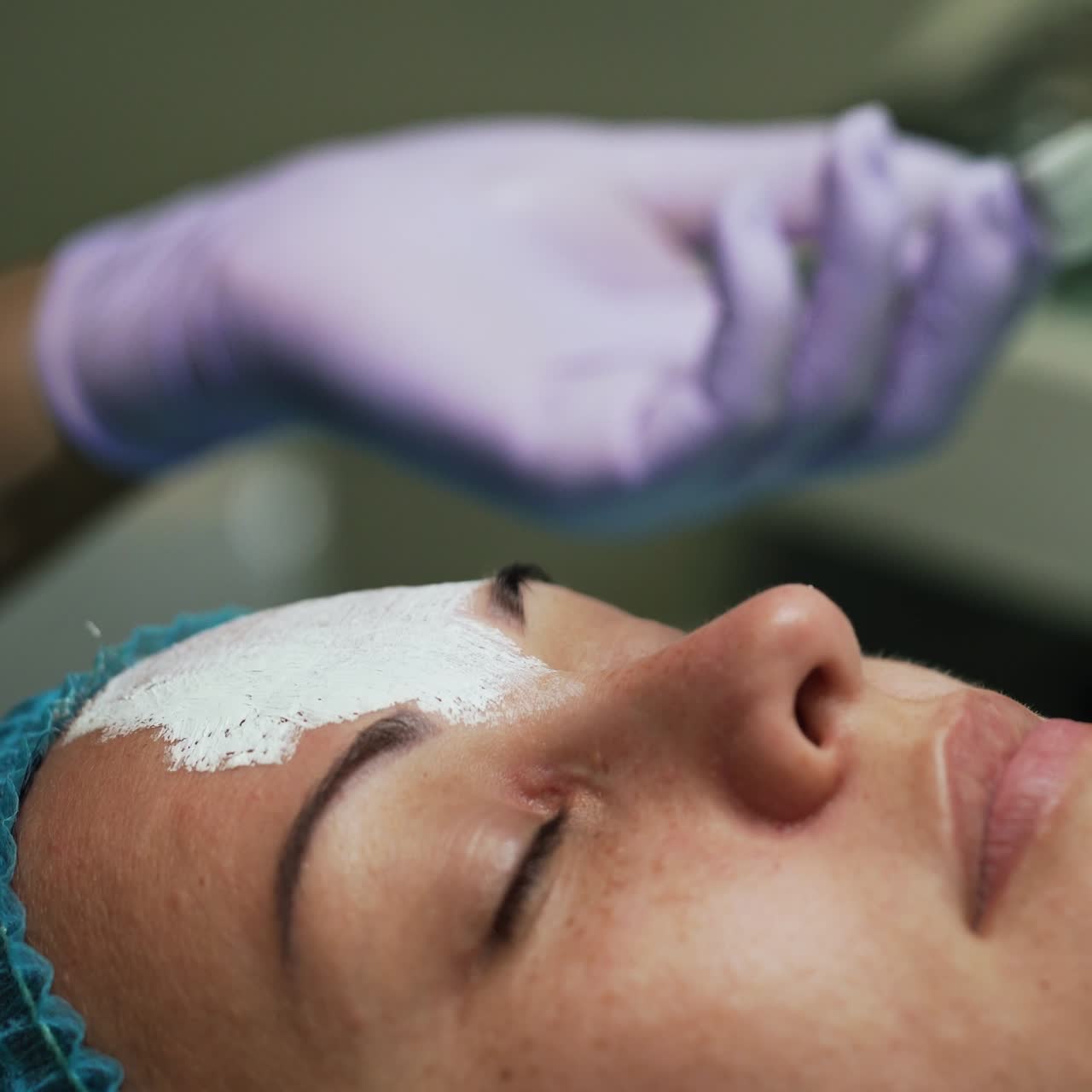 Beautician in latex gloves uses brush to cover client's face with mask. Rejuvenation procedures in beauty salon. Close up