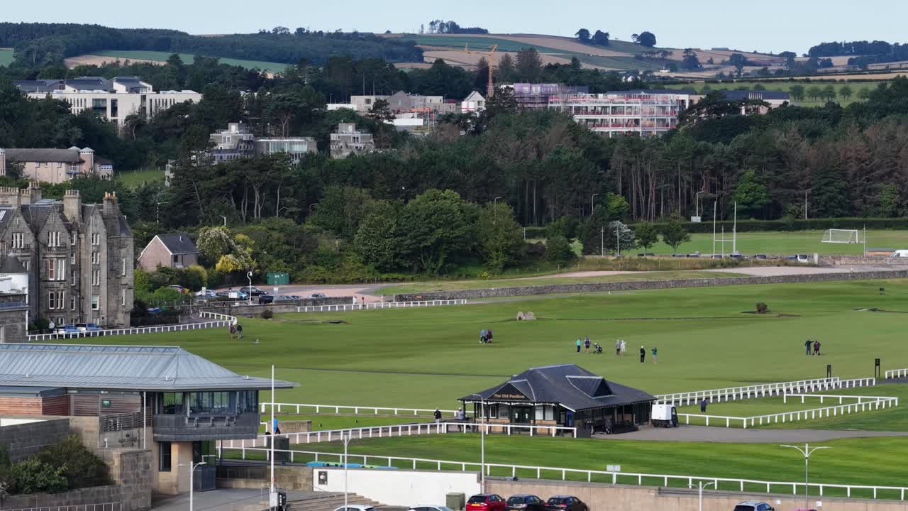 Golfers and tourists stroll across lush putting green near historic buildings, overcast daylight, wide shot