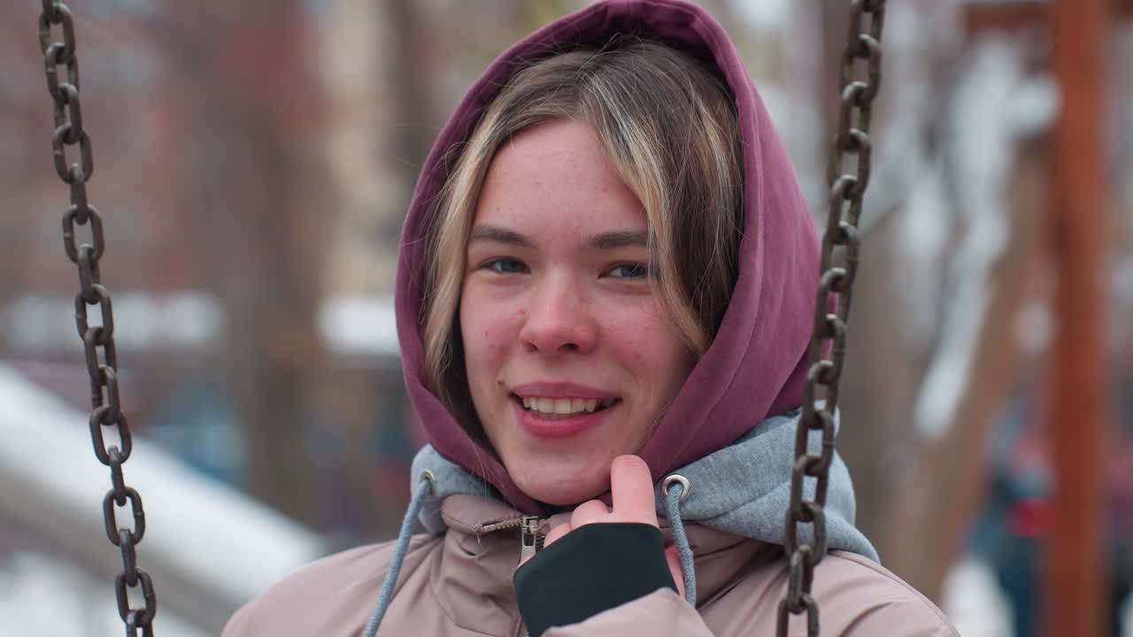 수줍어한 젊은 여성은 허디를 부드럽게 들고, 겨울 공원에서 스에 앉아, 따뜻한 재을 입고, 그녀는 부드러운 미소와 나무를 특징으로하는 흐릿한 배경으로 추운 날씨를 즐니다.