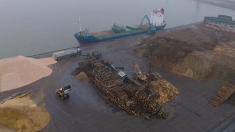 aerial establishing view of wood terminal crane loading timber into the cargo ship, port of liepaja , lumber log export, overcast day with fog and mist, birdseye drone shot moving forward