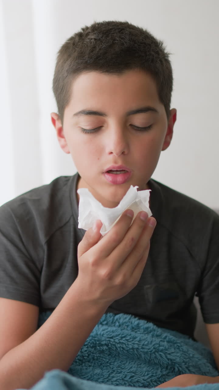 Sick boy sneezing into napkin covered with blanket on couch, wiping nose with tissue while looking tired, showing cold symptoms near window with soft light filtering in