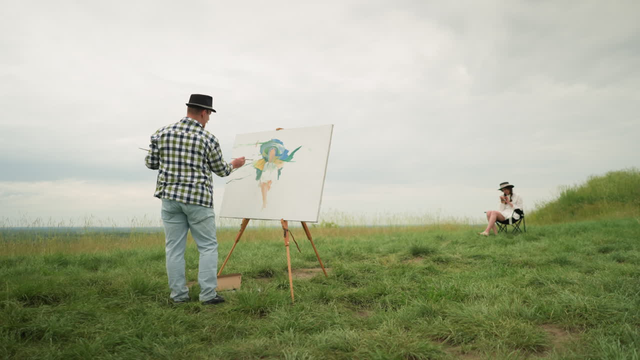 A craftsperson, wearing a hat and checkered shirt, is deeply focused on painting on a board in a lush grassy field under a cloudy sky. In front of him, a woman dressed in a white dress and hat sits