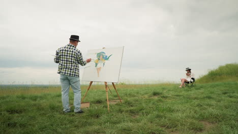 a craftsperson, wearing a hat and checkered shirt, is deeply focused on painting on a board in a lush grassy field under a cloudy sky. in front of him, a woman dressed in a white dress and hat sits